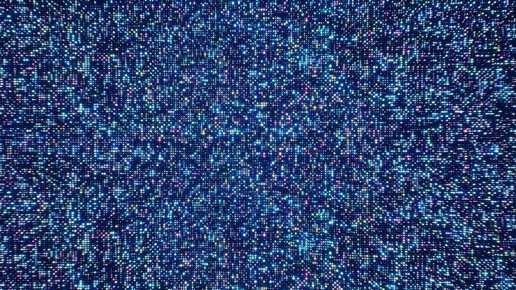 Un campo astratto di dense particelle di luce blu, viola e ciano riempie l'inquadratura con un effetto glitter luminoso, creando uno sfondo futuristico e dinamico.