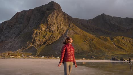 Zeitlupe,-Eine-Wunderschöne-Junge-Frau-In-Einer-Roten-Jacke-Bewundert-Die-Aussicht-Und-Lächelt-Am-Strand-Von-Flakstad-Vor-Einem-Wunderschönen-Goldenen-Sonnenuntergang,-Lofoten-Inseln,-Norwegen