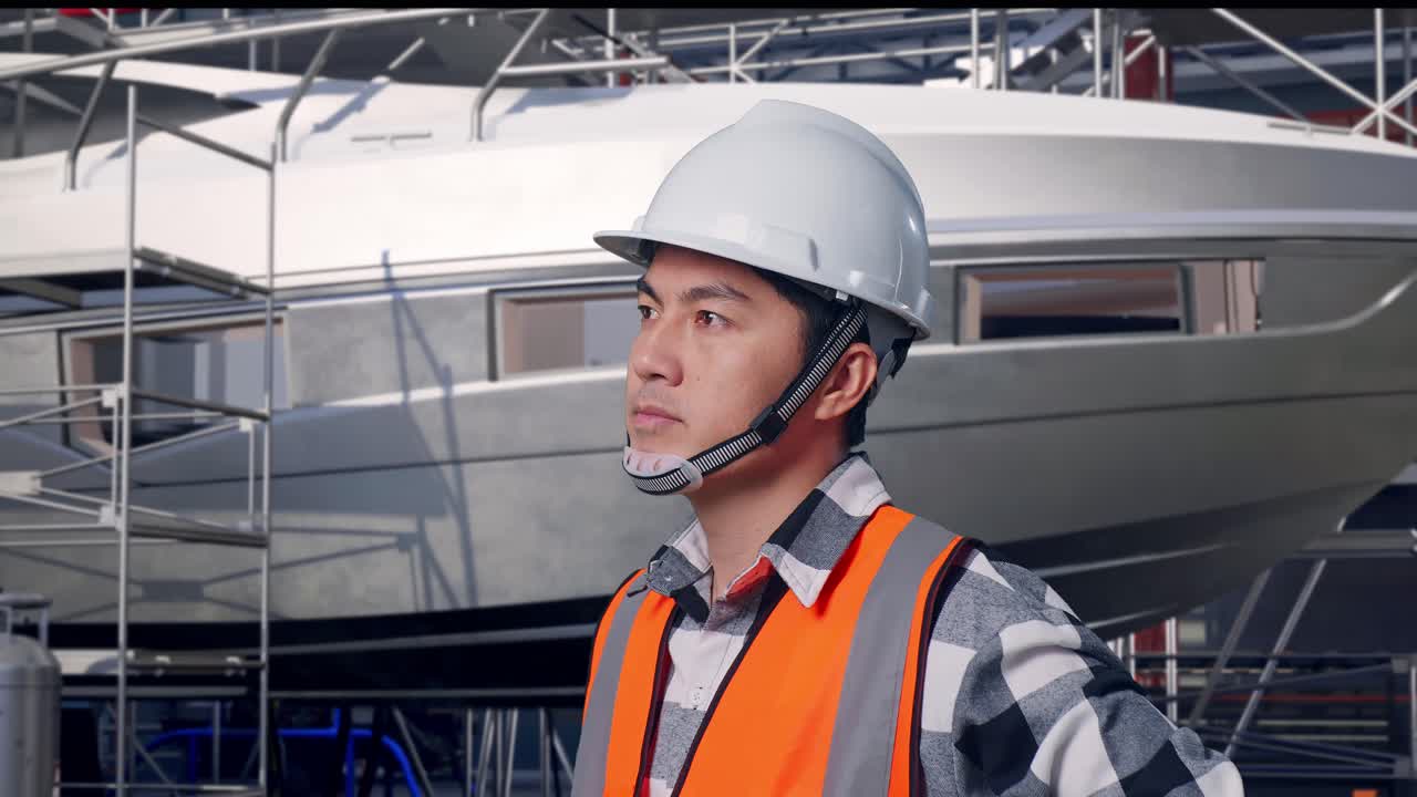 Primo piano laterale di un ingegnere asiatico che indossa un casco di sicurezza, si guarda intorno e sta in piedi a braccia conserte con la costruzione di yacht in un moderno cantiere navale