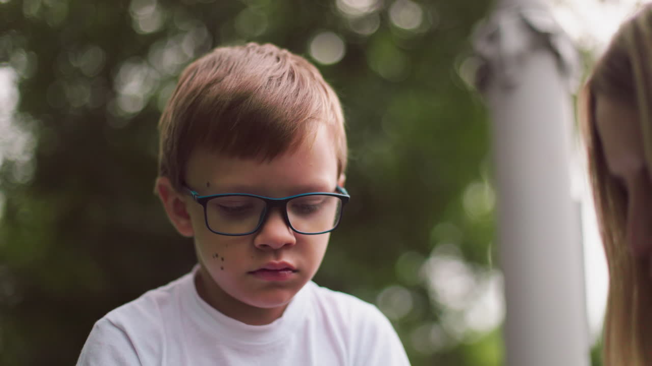 маленький мальчик в очках выглядит подавленным и задумчивым, глядя вниз с размытым фоном зелени и частичным взглядом на голову женщины