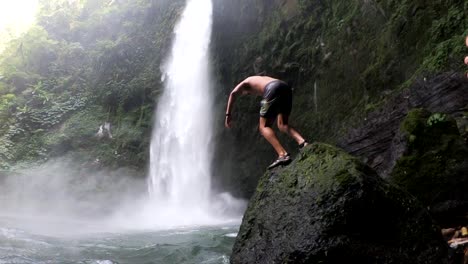 Slow-Motion-Shot-of-a-man-doing-a-front-flip-off-of-a-rock-and-into-the-swimming-hole-at-the-base-of-the-gushing-NungNung-Waterfall-in-Bali,-Indonesia