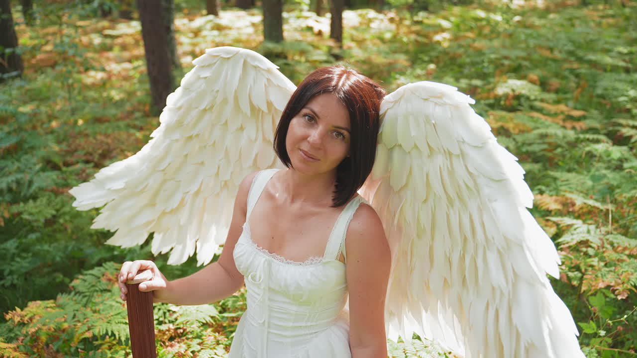 Lady dressed as angel with large white wings and wooden staff standing in bright green forest bathed in sunlight smiling softly with peaceful expression symbolizing hope