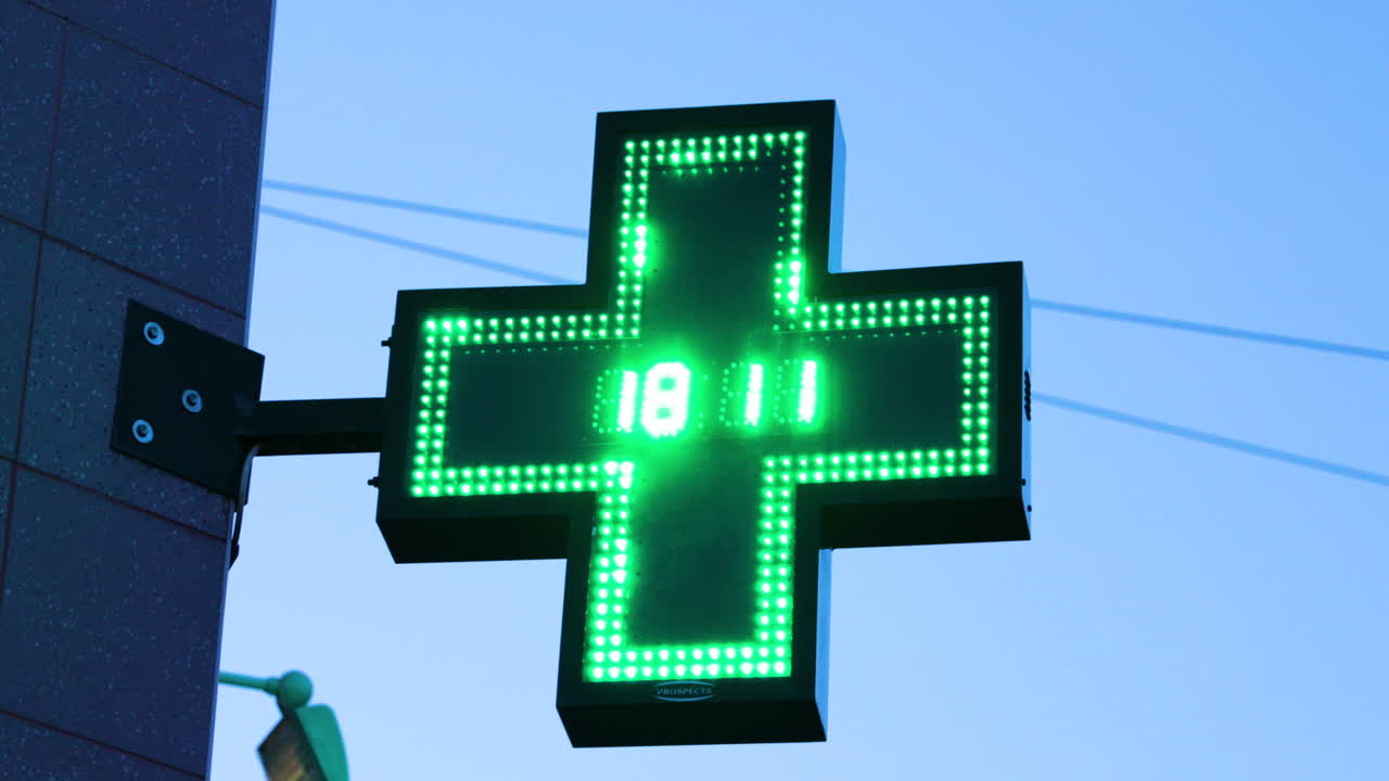 A pharmacy and illuminated international symbol hanging on a corner of a building with a twinkling clock in the middle of a green cross depicting a pharmacy caputred in 4k 60fps