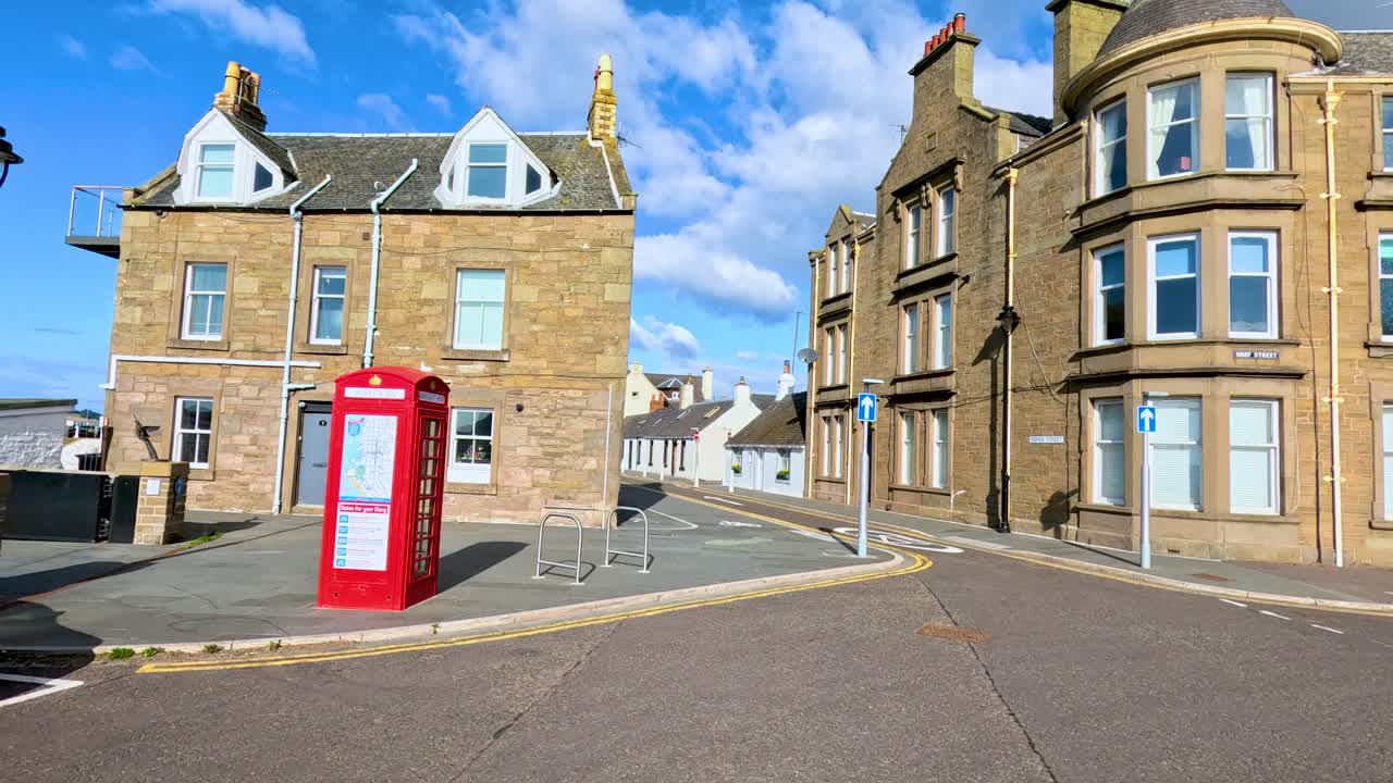 A bright daytime journey through quiet, historic streets near Broughty Ferry harbour, showcasing stone buildings, parked cars, and a classic red phone box. Smooth camera movement, natural light, and clear blue skies