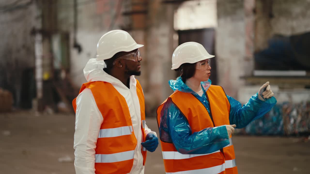 A man with Black skin in a white uniform together with his colleague a girl in an orange vest walk along the large hall of a waste processing plant and communicate with each other