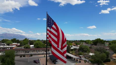 Amerikanische-Flagge-Weht-Im-Wind-An-Einem-Schönen-Sonnigen-Sommertag-Mit-Wolken-Und-Blauem-Himmel-Mit-Blick-Auf-Berge-Und-Einer-Kleinen-Stadtdrohne,-Die-Um-Den-Fahnenmast-Fliegt-–-In-4K-60fps