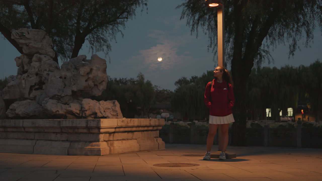Giovane donna in piazza sotto la luce della luna, con felpa rossa e skateboard vicino a un monumento in pietra sotto la calda luce di un lampione, in una notte urbana tranquilla, con una postura contemplativa e giochi di ombre tenui, in una solitudine cinematografica della città.