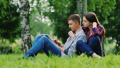 Young-Couple-Man-And-Woman-Sitting-On-The-Grass-In-The-Park-Enjoy-The-Tablet-Girl-Pressed-Against-Th