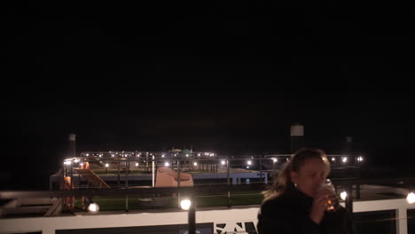 elegant woman in defocus savoring beverage while standing on nighttime terrace, overlooking illuminated urban skyline with serene, contemplative expression. filmed with vintage glass