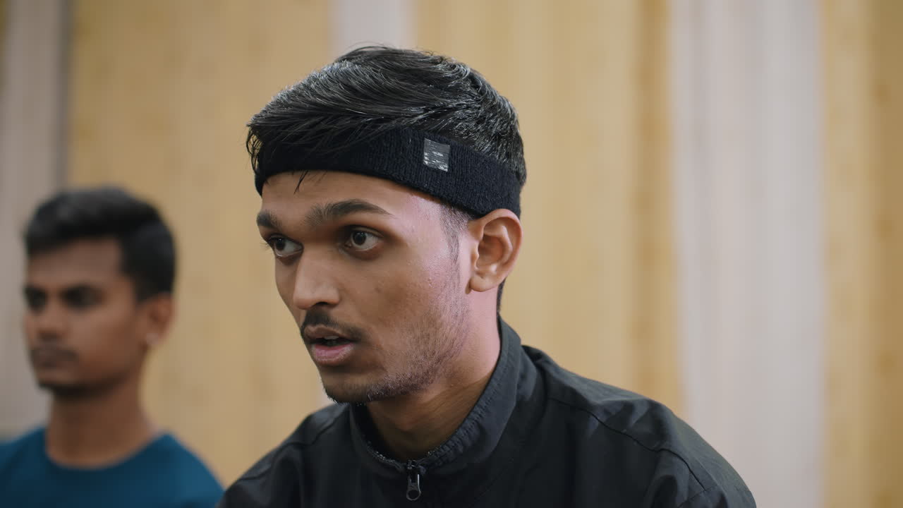 Close up of young Indian athlete wearing black headband and jacket concentrating during table tennis match as teammate in blue shirt stands in background