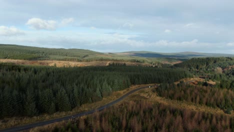 Drone-pull-back-revealing-vast-countryside-landscape-in-UK-with-2-cars-racing-down-single-track-gravel-road-on-sunny-but-cloudy-day