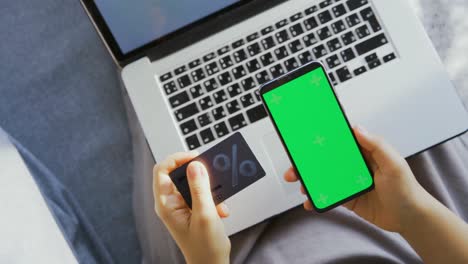 mujer sostiene una tarjeta de descuento y un teléfono móvil por encima de una computadora portátil