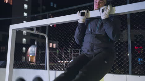 athlete gripping goal post performing pull-ups during a nighttime training session, showcasing strength and endurance with an illuminated urban backdrop