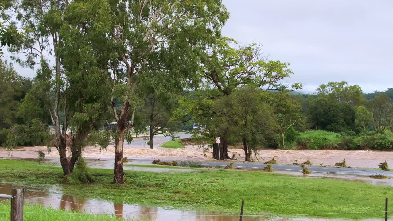 Wide shot of a river that burst its banks on the Gold Coast, Queensland, Australia. Fast-moving brown water rushes past green trees and grass, showing the power of floodwaters.
