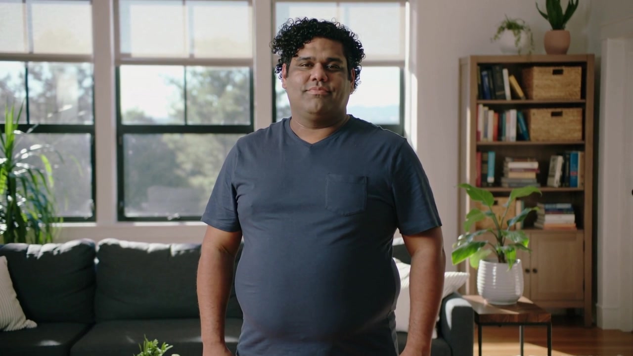 Kameraet starter, mand i marineblå T-shirt og beige bukser justerer holdning og smiler hjemme ved en sofa.