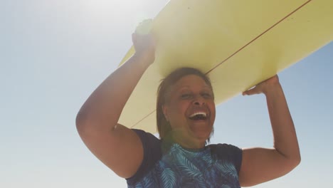 mujer afroamericana mayor feliz caminando con tabla de surf en una playa soleada