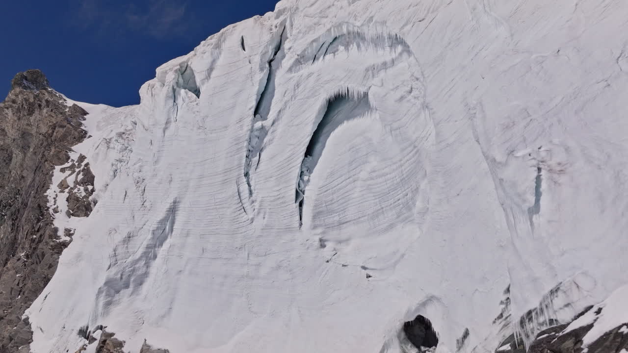 A powerful drone shot revealing massive overhanging seracs and towering ice cliffs in Saas-Fee, Switzerland. The frozen formations glisten under alpine light, showcasing the raw power of the glacier
