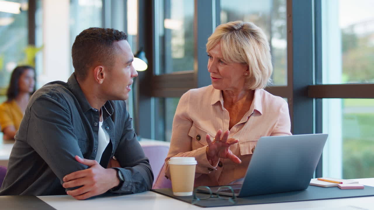 mujer de negocios madura que orienta a un colega más joven que trabaja en una computadora portátil en el escritorio