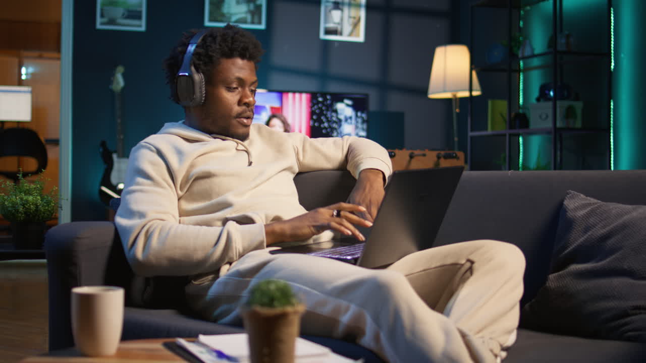 Black young adult attending a webinar on laptop from his living room, relaxing on his couch and listening on headset. Cheerful man multitasking in order to finish his daily tasks. Camera B.