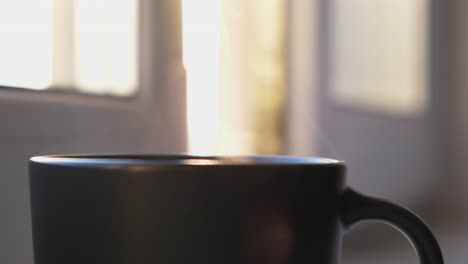 a cup of hot coffee or tea and the steam rising up in the air. concept. close up of black cup with hot beverage standing in front of the window at home on blurred background