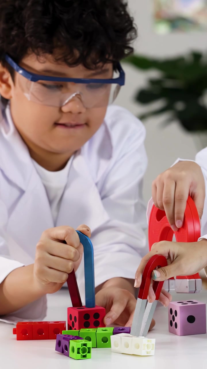 Elementary students wear safety goggles while testing magnetic forces on colorful blocks in classroom.