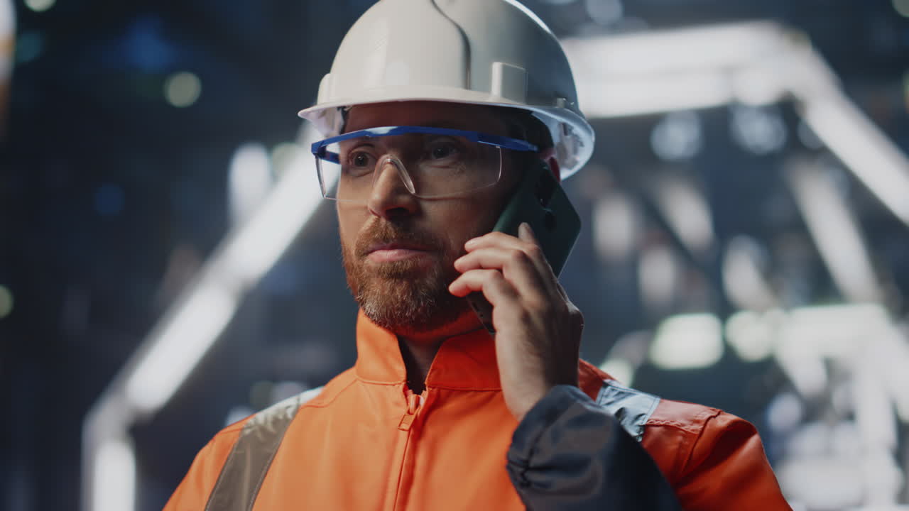 Operaio di fabbrica che parla al cellulare, in piedi in un impianto industriale, in primo piano.
