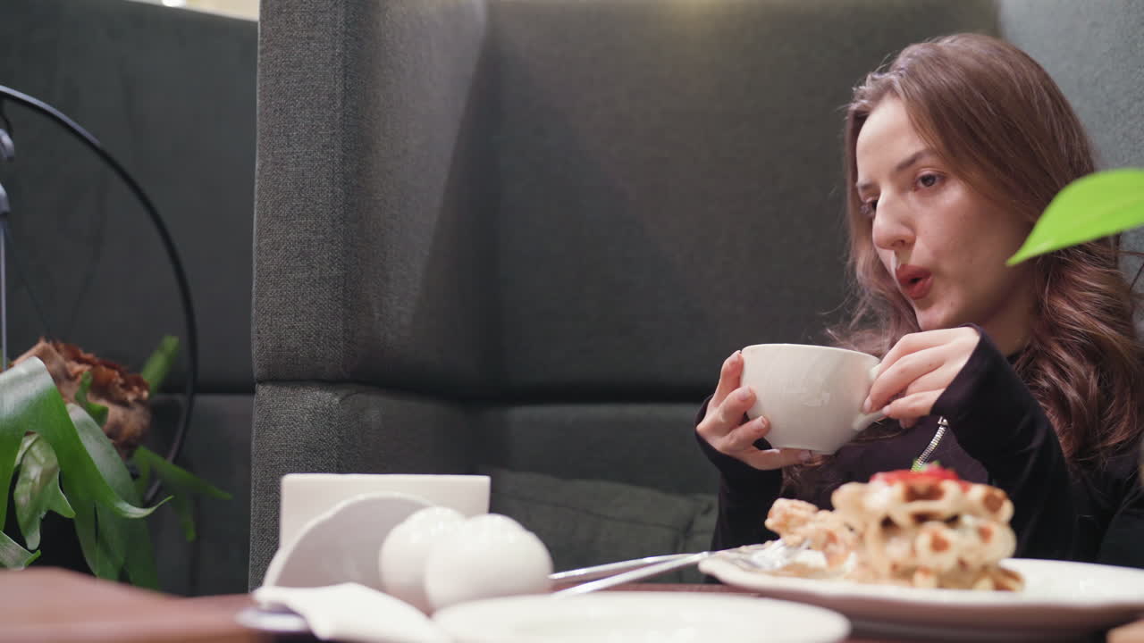 Lady with long wavy hair lifts white cup of hot latte, blowing softly to cool it down while seated at cozy booth with waffles, napkins, and warm ambient lighting in calm, relaxing setting
