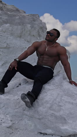 man with perfect body pumping up muscles and enjoying of the summer. multiracial man sitting at the sand. grey rocks background. vertical video