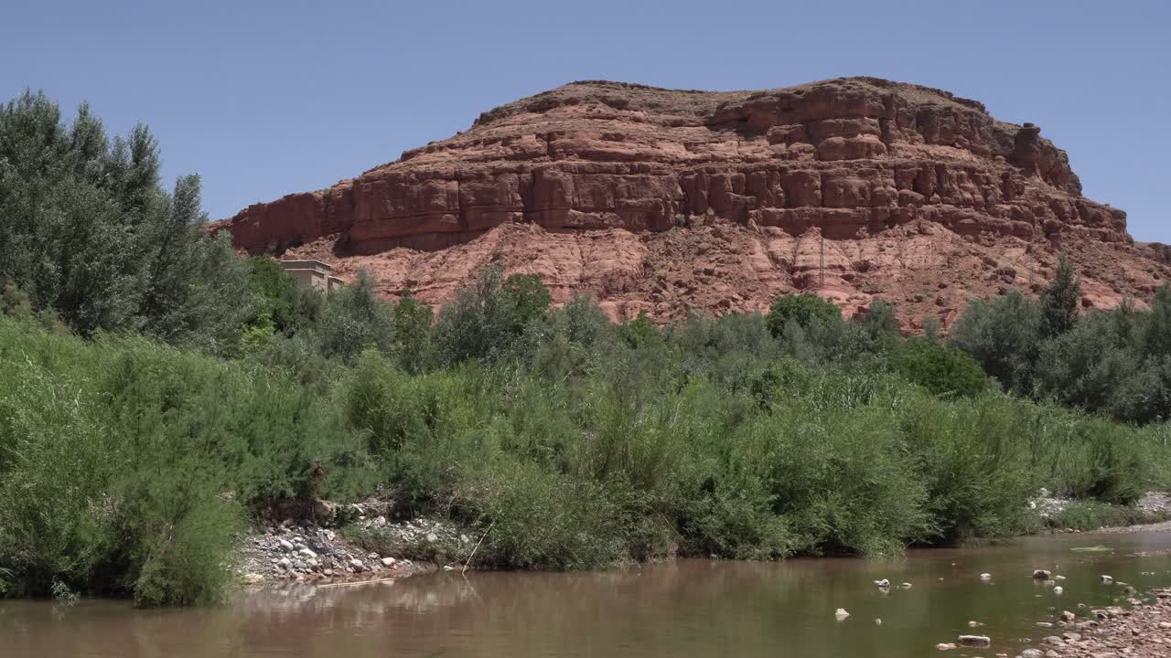 A desert mountain with no vegetation under a blue sky. In the foreground, there is a small river with lots of greenery on the other side