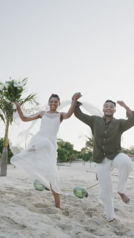 vertical video of happy diverse bride and groom holding hands jumping at beach wedding, slow motion