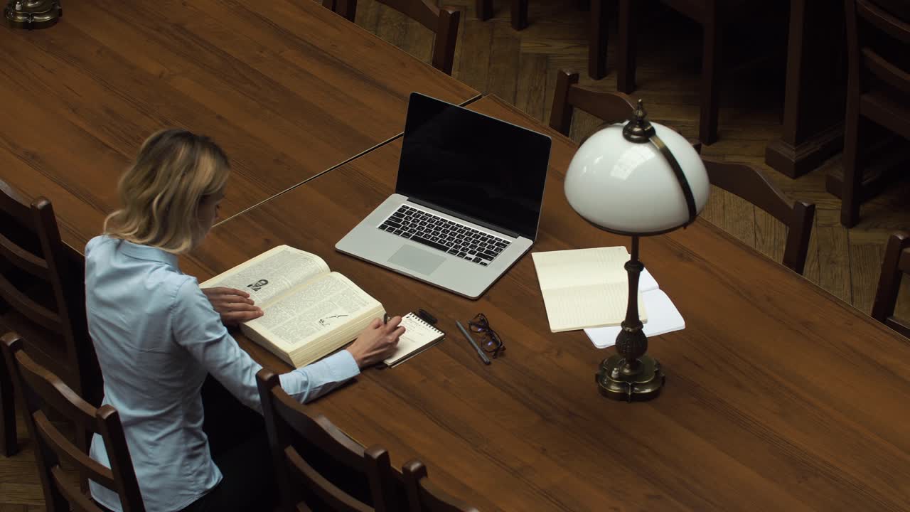 Woman reading a book with a laptop on the table