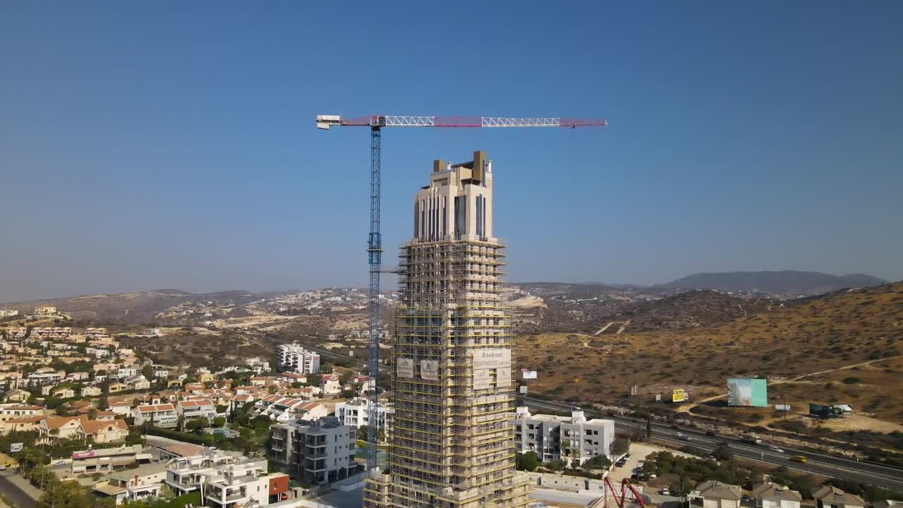 Drone footage captures a towering high-rise under construction in Sarandë, surrounded by hilly urban landscape. Bright daylight illuminates the structure and crane, accentuating architectural beauty