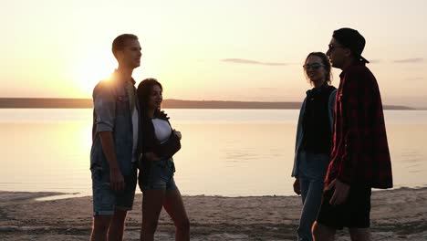 group of friends hanging together on the beach. sunset above the water. beautiful two couples. talking, gesturing. friendship concept