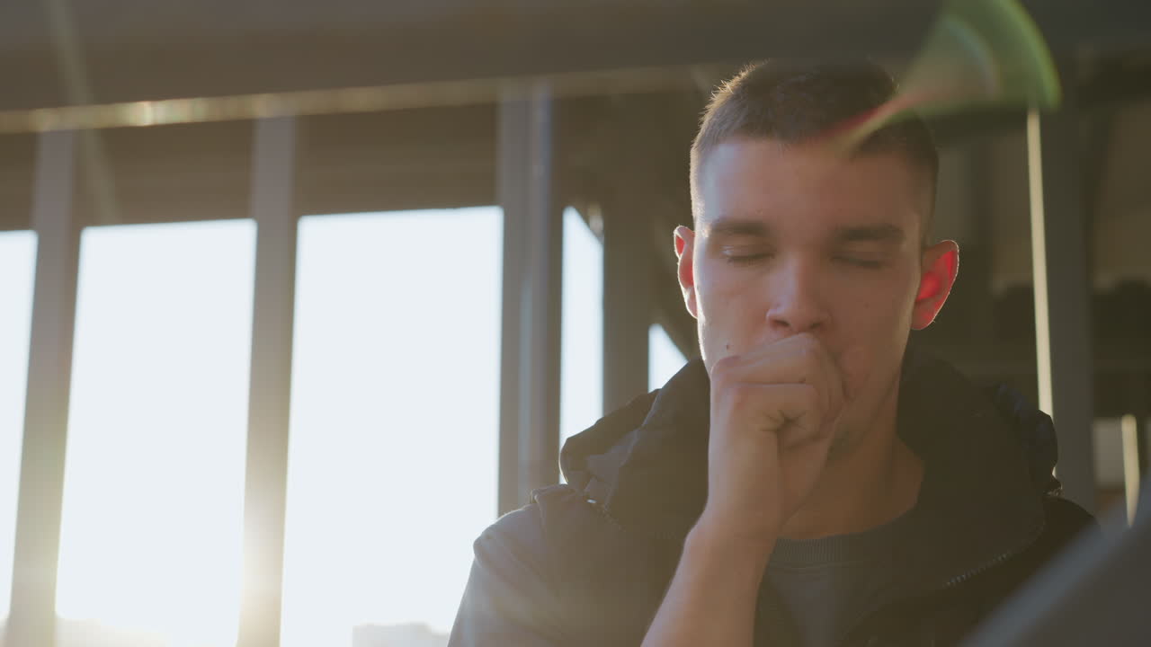 man in black padded vest standing outdoors holding medicine box in right hand, looking down with serious expression under bright warm sunlight near modern brick building with shadow lines