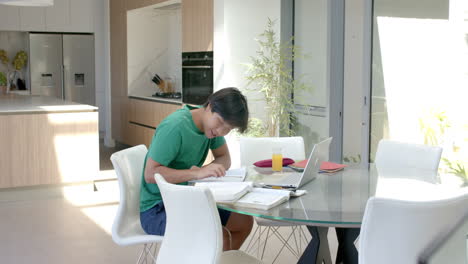 teenage asian boy studying on a laptop at a modern home kitchen table