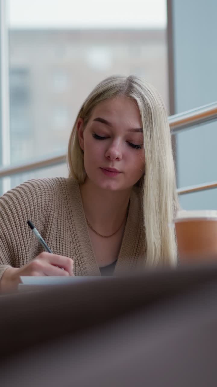 mujer joven escribiendo pensativa en el centro comercial con taza de café en la mesa de madera, vista parcial de objeto poco claro en el fondo, ambiente casual y pacífico