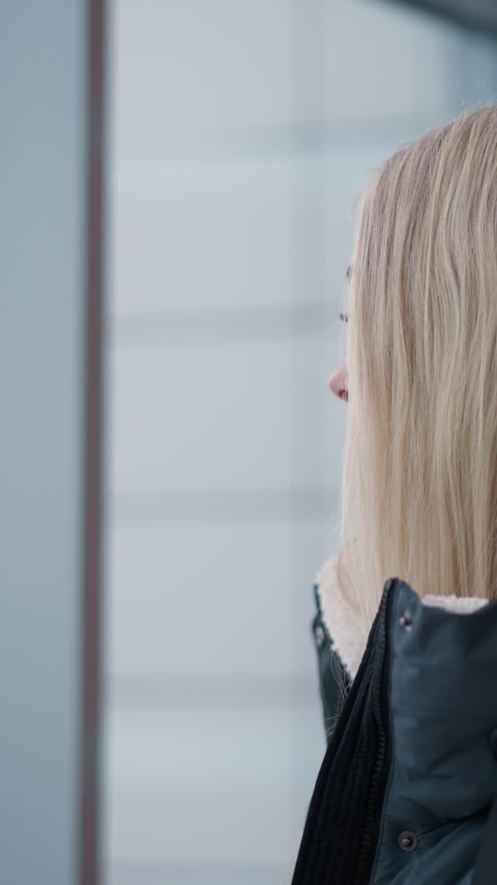 Signora con i capelli che le scendono sulla spalla, con uno sguardo contemplativo e uno sfondo urbano visibile attraverso la finestra di vetro, un momento di serenità e riflessione.