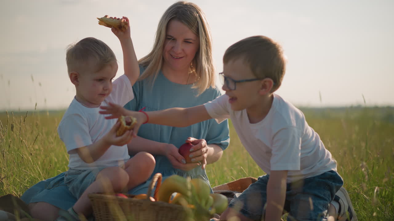 una madre con un vestido azul se sienta en la hierba con sus dos hijos, ambos con camisetas blancas. el hijo menor está hablando alegremente mientras sostiene un bocadillo, y el hijo mayor se acerca para recoger uno de él
