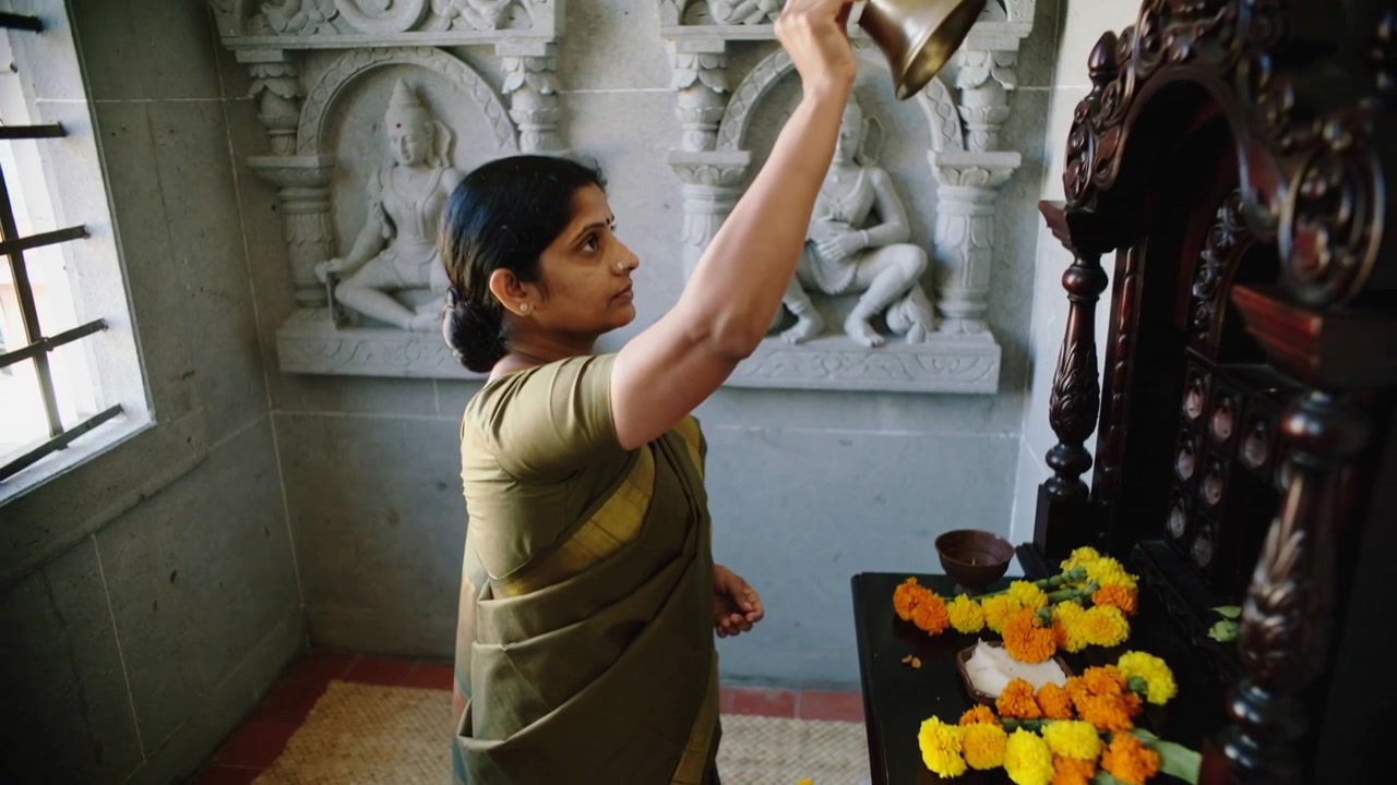 Raising brass bell, woman in olive-green sari ringing during prayer at small shrine, placing bell