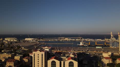 vuelo de drones sobre un complejo de edificios hasta el puerto con grúas de carga y barcos al atardecer y el horizonte con el mar, haifa, israel