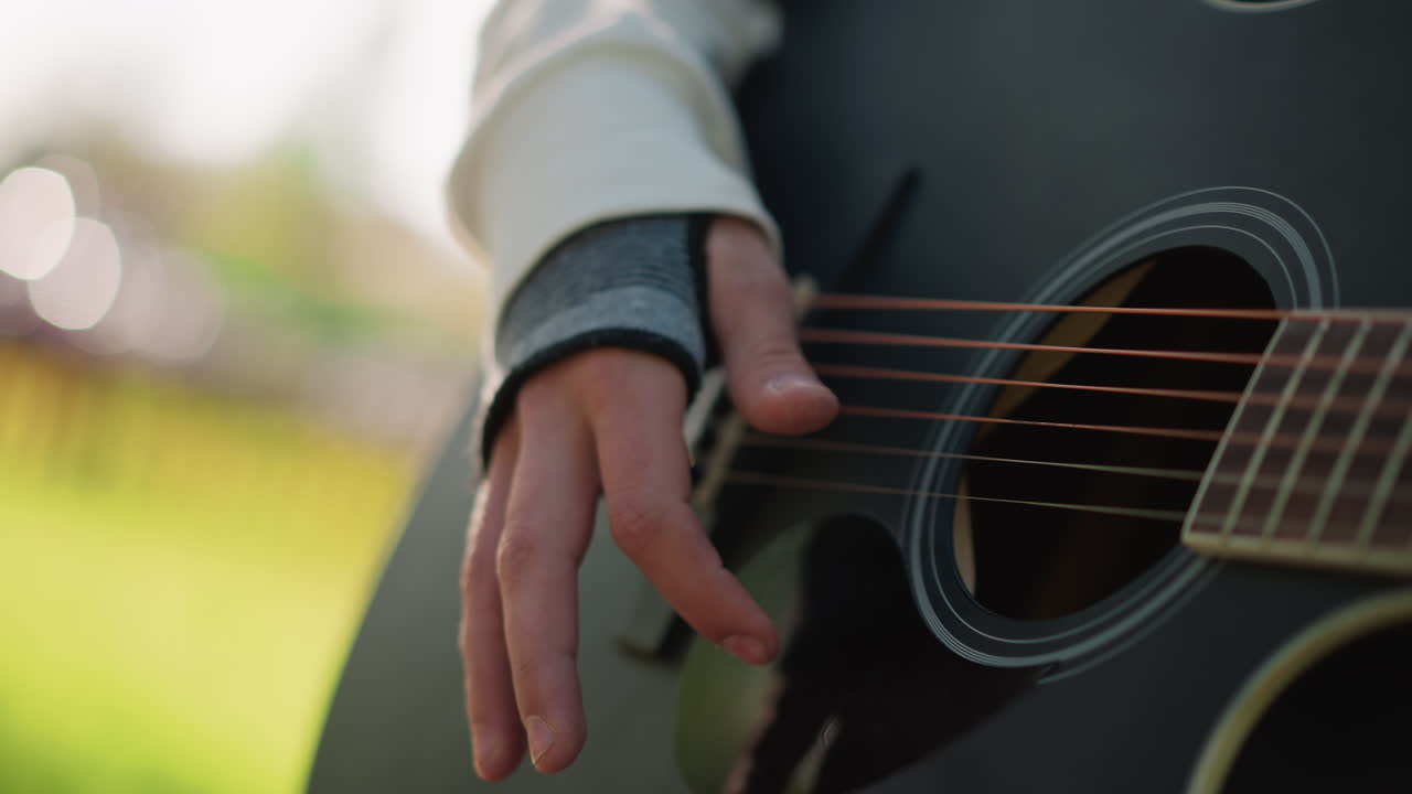 primo piano di mani che pizzicano una chitarra acustica all'aperto, chitarra nera consumata con corde visibili, bokeh delicato e luce solare dorata sull'erba, manica di maglione casual, fingerstyle picking, melodia lenta e riflessiva