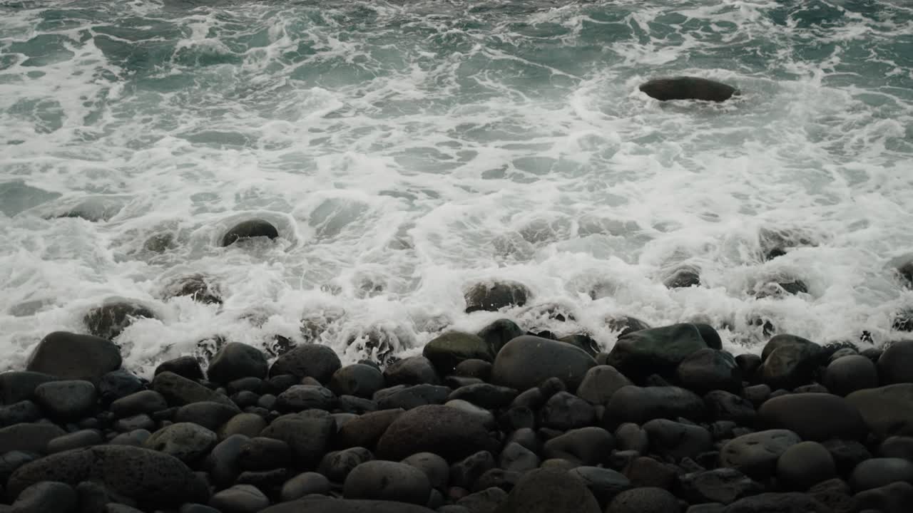 Waves roll over smooth dark stones along Madeira’s coast, creating a rhythmic pattern of motion and foam beneath overcast skies.