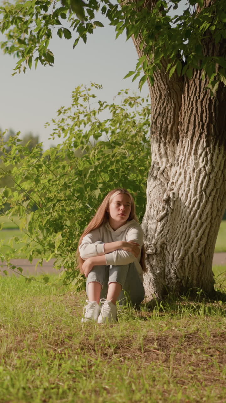 mujer se sienta en el suelo cubierto de hierba bajo un árbol con expresión pensativa, cruzando los brazos sobre las piernas, la luz del sol se refleja en su cara mientras mira hacia la derecha, el fondo incluye postes y estadio