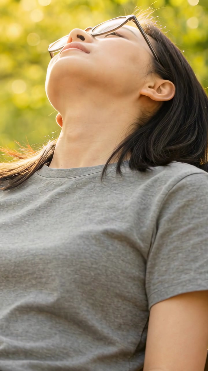 Vertical video: Tilting Asian woman leaning back on park bench in sun, wearing grey tee and glasses