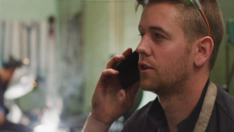 Caucasian-male-factory-worker-at-a-factory-standing-in-a-workshop,-talking-on-a-smartphone