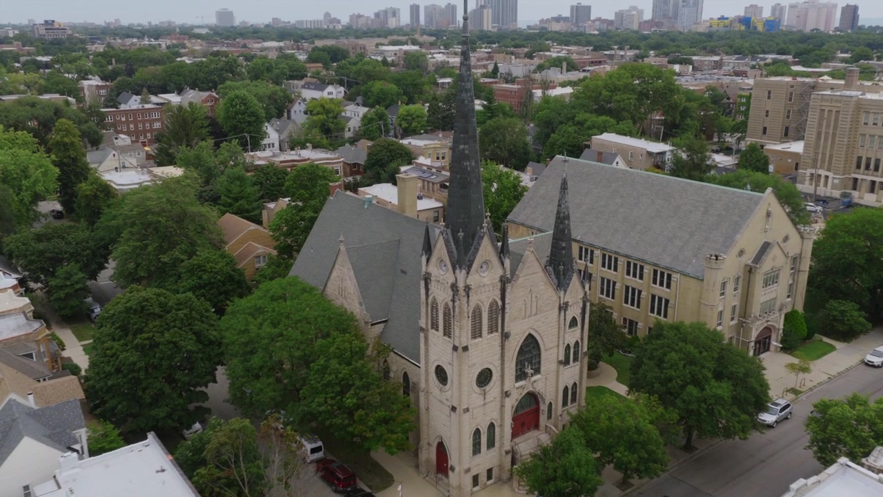 Imagem aérea de drone de um bairro de Chicago centrada em uma igreja e escola góticas, com torres altas e o horizonte à beira do lago ao fundo