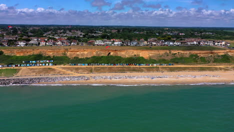 Luftaufnahme-Der-Küste-Mit-Strandhütten,-Gleitschirmfliegern-Und-Küstenstadt-An-Einem-Sonnigen-Tag-In-Großbritannien,-4k