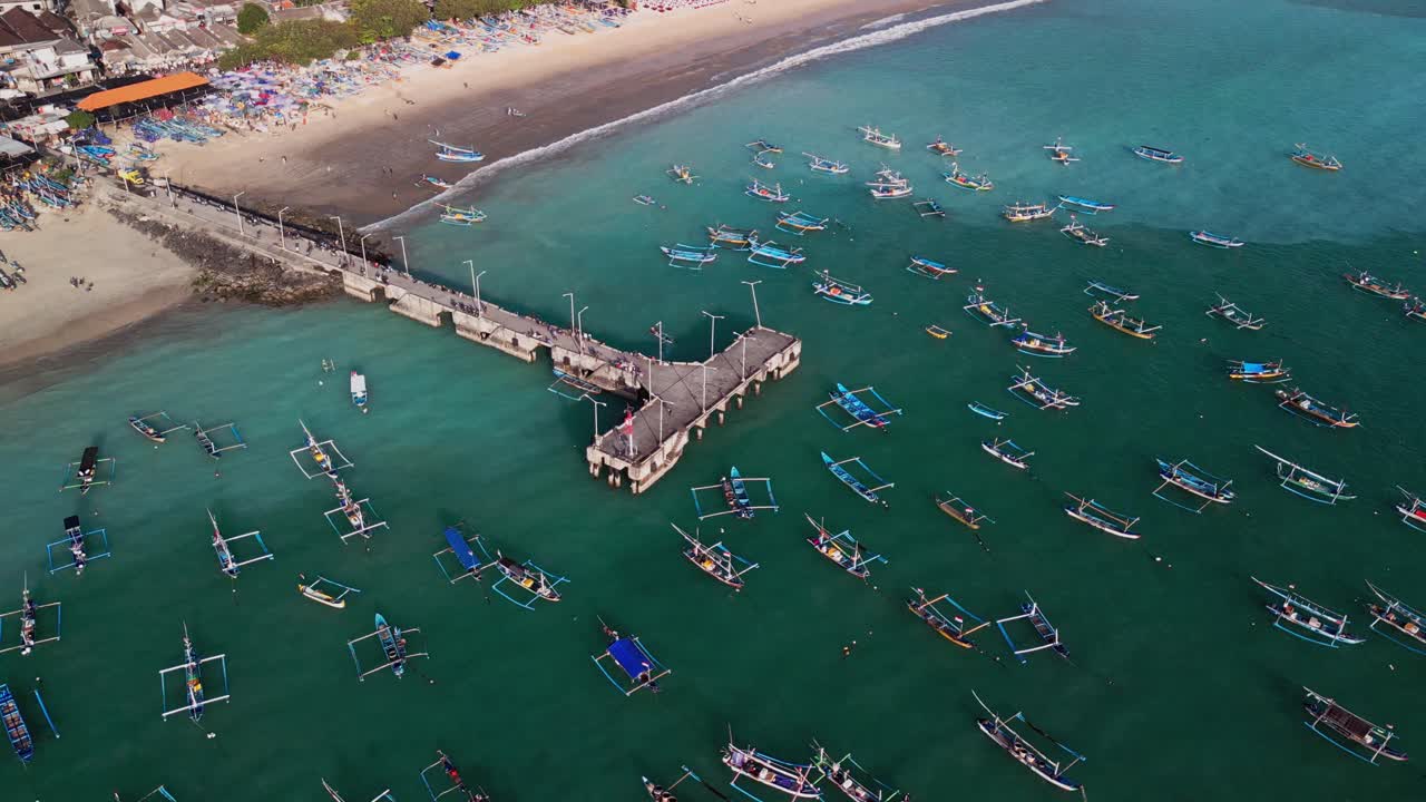 Unique aerial view showcases Jimbaran harbor where fishing boats gather in clear lagoon, beachfront settlement and pier walkway, highlighting the balance of tradition and modern life in Bali