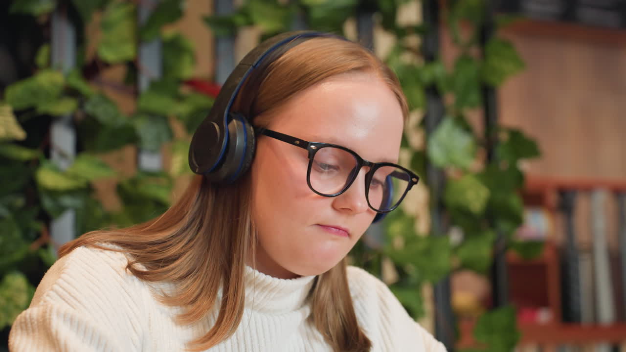 Close up of girl wearing black glasses and headphones gently dancing to music, enjoying moment with subtle smile, soft blur background with green indoor plants and warm ambient light