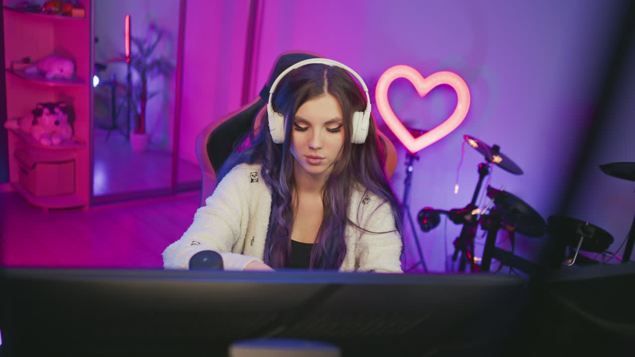 Young woman focused on work in colorful studio, pauses briefly to reach for popcorn before continuing task on computer, seated in modern gaming chair with glowing heart light in background